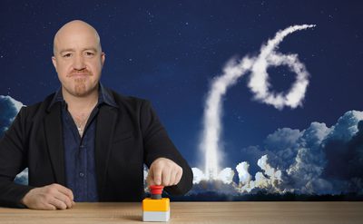 Person seated at a table pressing a large red button, with a dramatic night sky in the background showing a rocket launch leaving a smoke trail shaped like the number six.
