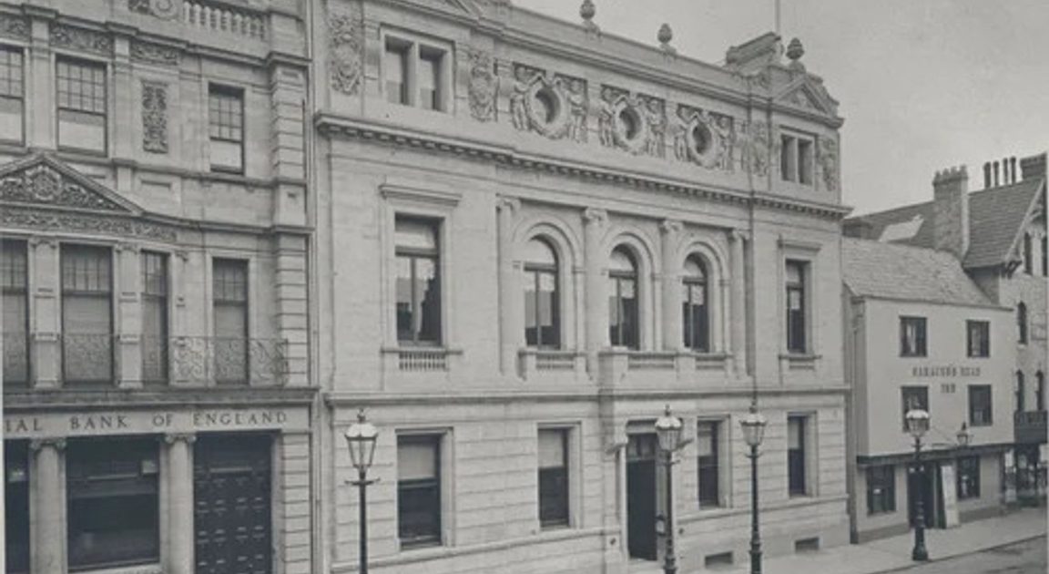Street view featuring classical architecture with ornate buildings including the National Bank of England on the left and Freemasons Hall on the right, with street lamps lining an empty sidewalk.