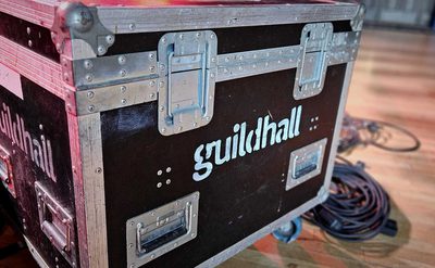 Large black metal-reinforced equipment case on wheels with 'guildhall' stenciled in white, placed on a wooden floor with coiled cables and other gear in the background.
