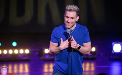 A man with short spikey blonde hair wearing a purple tshirt stands holding a microphone and smiling towards the audience.