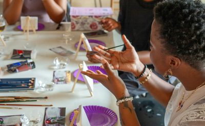 A group of people seated around a table participating in an art activity, with easels, canvases, and painting supplies.