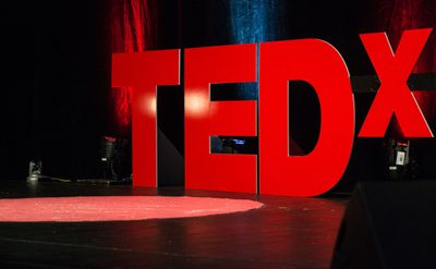 A photo taken from the side of a stage with the logo for TED x in large red letters at the back of the stage, there is a spotlight on the floor of the stage.