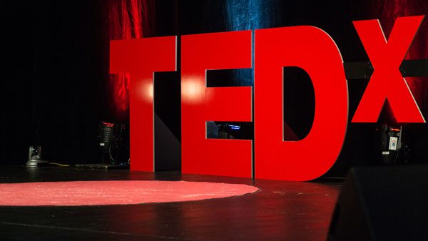 A photo taken from the side of a stage with the logo for TED x in large red letters at the back of the stage, there is a spotlight on the floor of the stage.