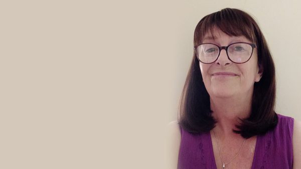 A lady with shoulder length brown hair, wearing black glasses and a purple top smiles at the camera.