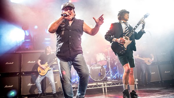 Live performance by a rock band on stage with five members: a central vocalist, two guitarists, a bassist, and a drummer. The guitarist on the right wears a schoolboy outfit and plays an electric guitar. The stage features smoke, colorful lights, and stacked Marshall amplifiers.