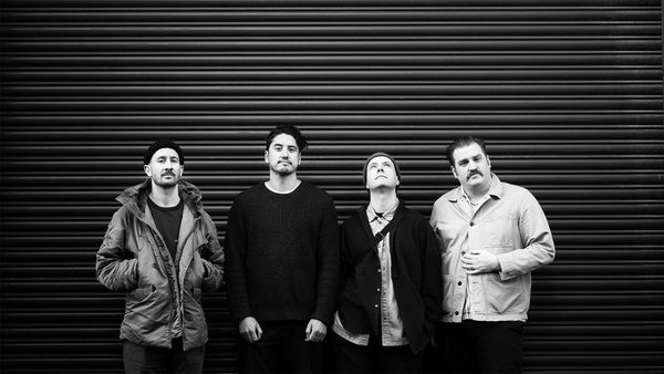 Black and white photo of four individuals in casual clothing, including jackets and sweaters, standing in front of a corrugated metal wall.