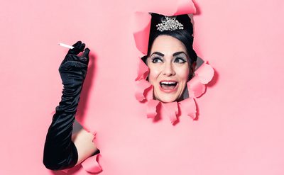 Person partially visible through a hole in a pink background, wearing a black outfit and long black gloves, with one arm extended holding a cigarette