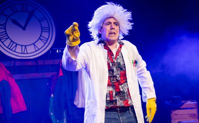 Person wearing a white lab coat, patterned shirt, and yellow gloves, pointing forward on a stage with a large clock in the background and colourful costumes hanging behind.