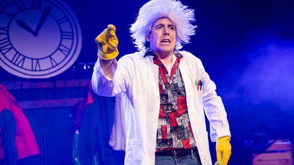 Person wearing a white lab coat, patterned shirt, and yellow gloves, pointing forward on a stage with a large clock in the background and colourful costumes hanging behind.