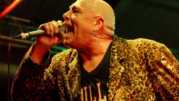 A bald man wearing a leopard print coat sings into a microphone.