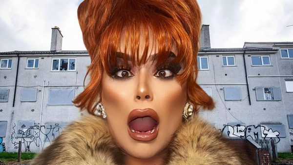 A person with red hair and a fur collar standing in front of grey apartment buildings with graffiti on the walls.
