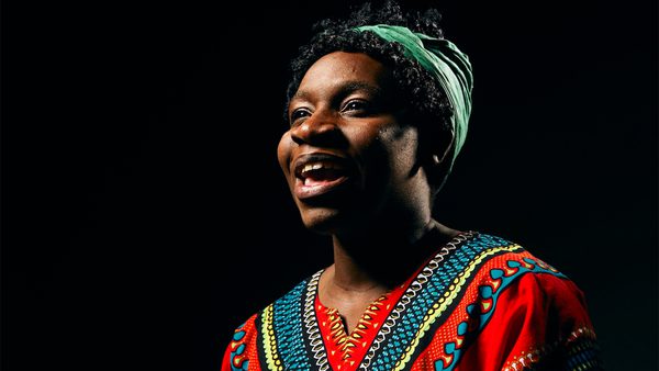 Person in colourful patterned clothing and green head wrap, lit against a dark background.