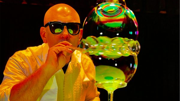 Person in a white shirt blowing a large, multi-layered bubble that reflects vibrant colours and light.