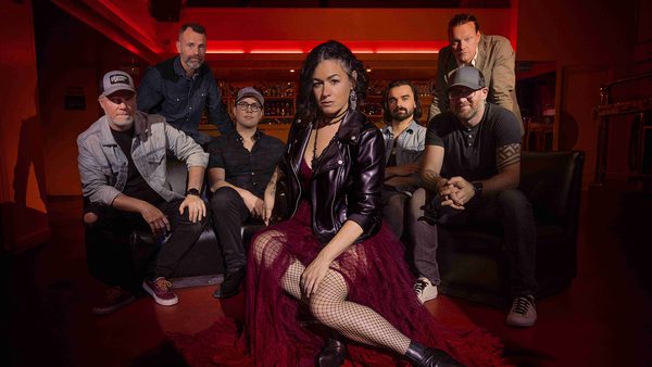 Six people posing in a dimly lit bar or lounge; central figure seated on a red carpet wearing a leather jacket, red dress, fishnet stockings, and black boots. Others are casually dressed with faces obscured.