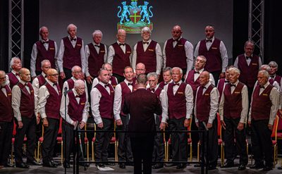 A male choir performing on stage, dressed in white shirts, maroon vests, and black bow ties, with a conductor in front and a crest featuring two lions and a cross in the background.
