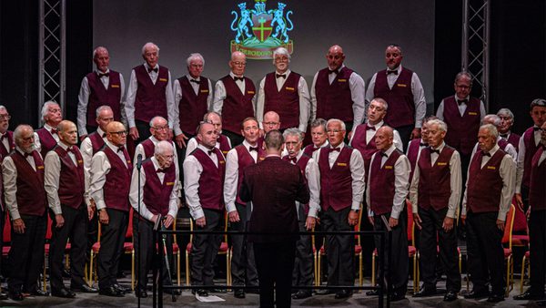 A male choir performing on stage, dressed in white shirts, maroon vests, and black bow ties, with a conductor in front and a crest featuring two lions and a cross in the background.