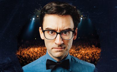 A man with dark hair and black glasses wearing a blue shirt with a black bow tie, frowns at the camera.