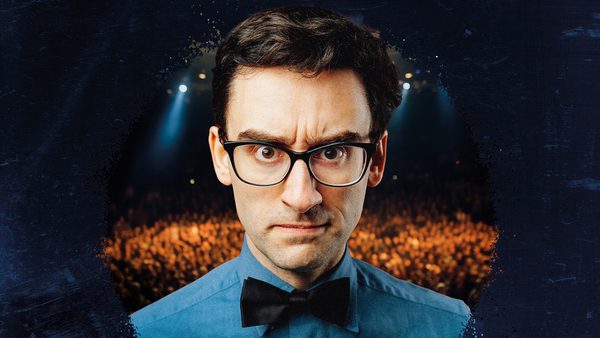 A man with dark hair and black glasses wearing a blue shirt with a black bow tie, frowns at the camera.