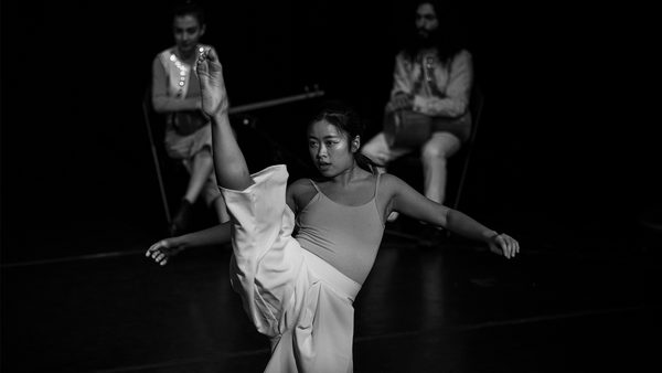 A person performing a high kick dance move on stage, wearing a sleeveless top and loose pants, with two other individuals seated in the background. All faces are obscured.