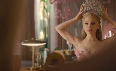 A person wearing a sparkling pink strapless gown is holding an ornate silver crown above their head in an elegant room with soft lighting, a vintage desk lamp, and decorative floral elements in the background.