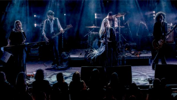 Band with five members performing on a stage illuminated by blue and purple lighting, creating a moody atmosphere; audience visible in front of the stage.