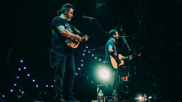 Two musicians performing on stage with guitars, standing in front of microphones. The background is dark with visible stage lights, and water bottles are placed near their feet.