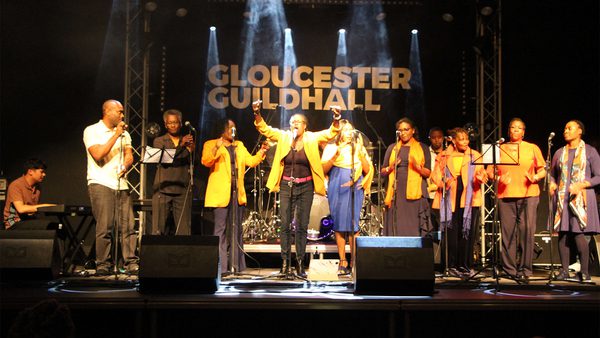 Group of performers on stage at Gloucester Guildhall, with some holding microphones and others standing in front of music stands under spotlights. The backdrop displays the text 'GLOUCESTER GUILDHALL'