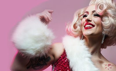 Person in a red sequined outfit with white fur trim, wearing a curly blonde wig. They have a tattoo visible on their upper arm. The background is pink.