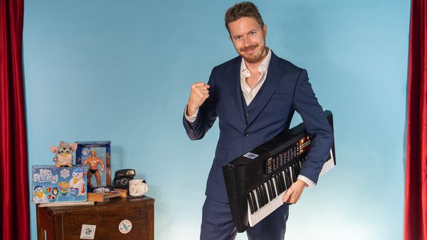A person in a dark blue suit stands in front of a light blue backdrop with red curtains, holding a keyboard instrument and raising one fist. To the left, a wooden table displays toys, books, a stuffed animal, an action figure, and board games or puzzles.