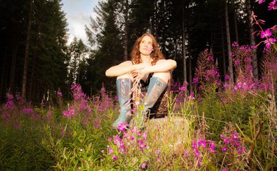 A photo of a woman sitting in a field of purple flowers surrounding her and tall trees behind her. She is wearing blue knee-high boots and a black, lace, knee length catsuit and is smiling to herself. She holds one of the purple flowers in her hand.