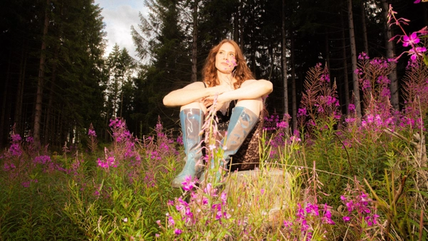 A photo of a woman sitting in a field of purple flowers surrounding her and tall trees behind her. She is wearing blue knee-high boots and a black, lace, knee length catsuit and is smiling to herself. She holds one of the purple flowers in her hand.