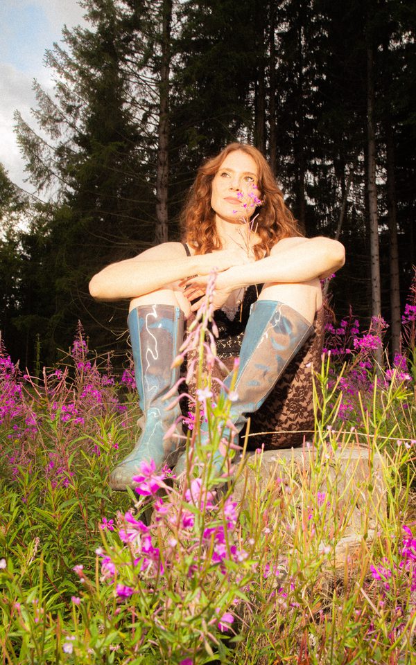 A photo of a woman sitting in a field of purple flowers surrounding her and tall trees behind her. She is wearing blue knee-high boots and a black lace, knee length jumpsuit and is smiling. She holds one of the purple flowers in her hand.