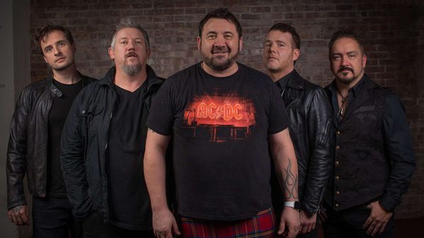 Five people standing in front of a brick wall with faces obscured; the central figure wears an AC/DC t-shirt and red plaid pants, others are dressed in dark clothing including leather jackets and vests.