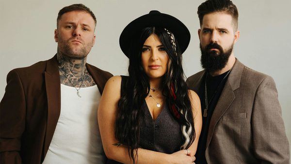 Three people standing close together. The person on the left wears a brown jacket over a white shirt with visible neck tattoos. The middle person has long braided hair, a black hat, and a dark sleeveless top. The person on the right wears a brown jacket over a dark shirt and has facial hair.