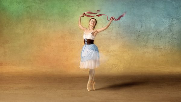 A ballet dancer performing on pointe, wearing a white and light blue dress with a brown belt, and holding red ribbons in both hands. The background features a gradient of green, yellow, and blue.