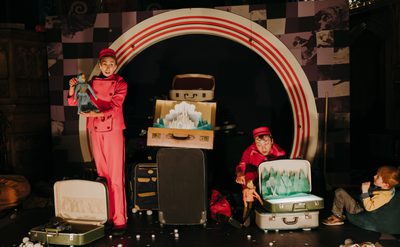 Two performers, a man and a woman in red jumpsuits and hats, are on stage with suitcases. The woman on the left holds a girl puppet dressed in blue, and the man on the right holds a boy puppet and looks at an open suitcase with a paper cutout of a castle inside. A child is seated on the far right, watching the performance.