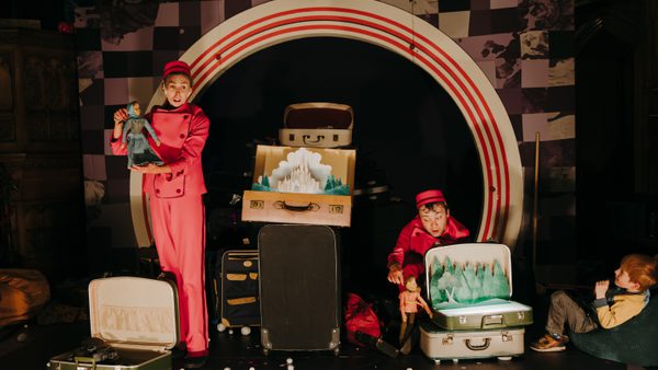 Two performers, a man and a woman in red jumpsuits and hats, are on stage with suitcases. The woman on the left holds a girl puppet dressed in blue, and the man on the right holds a boy puppet and looks at an open suitcase with a paper cutout of a castle inside. A child is seated on the far right, watching the performance.