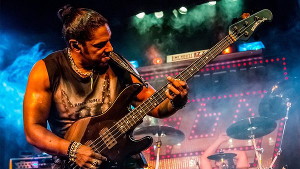 A musician playing an electric bass guitar on stage, wearing a sleeveless 'ROCK ROLL' shirt, bracelets, and a necklace. Stage lights, smoke effects, and a drummer are visible in the background.