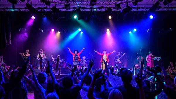 Lively concert scene with a band performing on a brightly lit stage featuring purple, blue, green, and yellow spotlights. Musicians are playing guitars and keyboards, while the audience in the foreground raises their hands in enjoyment.