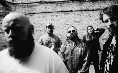 Black and white photo of five people standing in front of a weathered brick wall. The group is casually dressed, wearing graphic t-shirts, jackets, and caps, with some individuals positioned closer to the camera and others further back.