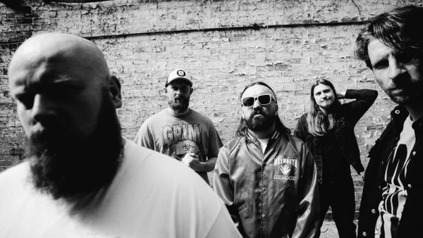 Black and white photo of five people standing in front of a weathered brick wall. The group is casually dressed, wearing graphic t-shirts, jackets, and caps, with some individuals positioned closer to the camera and others further back.