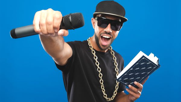 A person wearing a black t-shirt, gold chain necklace, and black cap, holding a microphone in one hand and an open book in the other. The face is blurred and the background is blue.