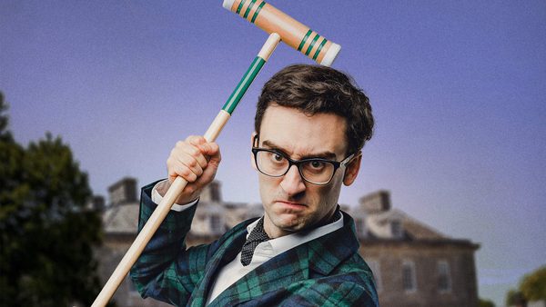 Person in a green and blue plaid jacket holding a croquet mallet above their head, standing in front of a large historic building with chimneys and surrounded by greenery under a clear blue sky.