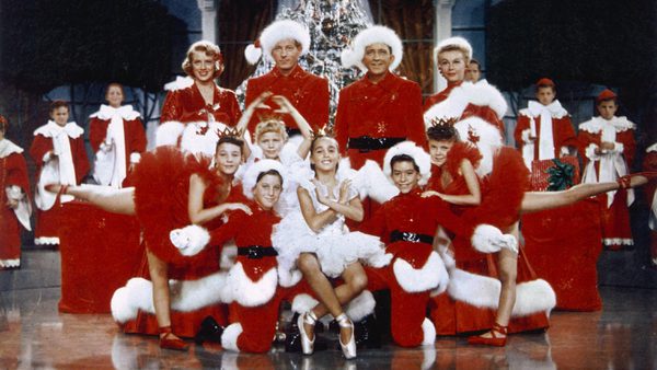 A group of people stand on a stage dressed in red and white outfits in front of a Christmas tree.