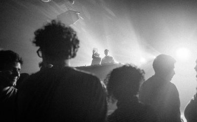 A black and white photo taken from a crowd, silhouettes of the backs of the audience in the crowd look ahead at two performers on stage staging behind DJ decks with bright lights behind them.