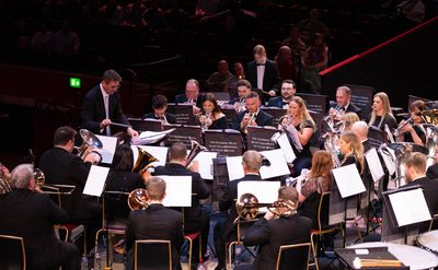 A photo of a brass band performing on stage, the musicians are sat in playing their instruments and looking at their sheets of music. The conductor is stood at the front of the band, in the background there is an audience sat in darkness watching the band.