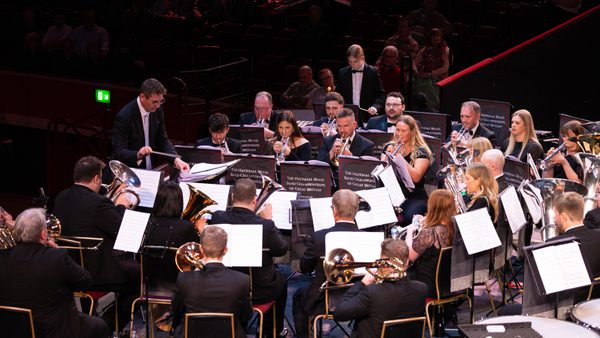 A photo of a brass band performing on stage, the musicians are sat in playing their instruments and looking at their sheets of music. The conductor is stood at the front of the band, in the background there is an audience sat in darkness watching the band.