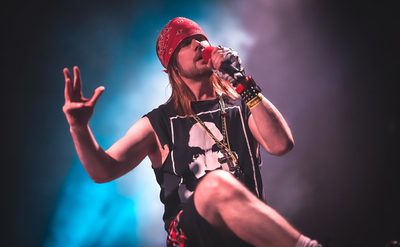 A man stands on stage holding a microphone, he is wearing a red headband. a black sleeveless t-shirt and shorts.