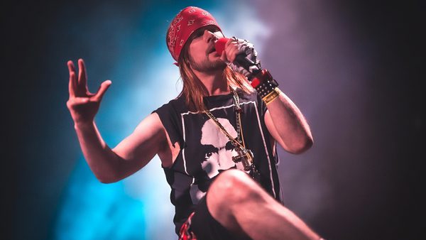 A man stands on stage holding a microphone, he is wearing a red headband. a black sleeveless t-shirt and shorts.