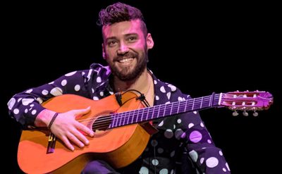 A man with dark hair wearing a black shirt with white spots on it, sits smiling holding an acoustic guitar.